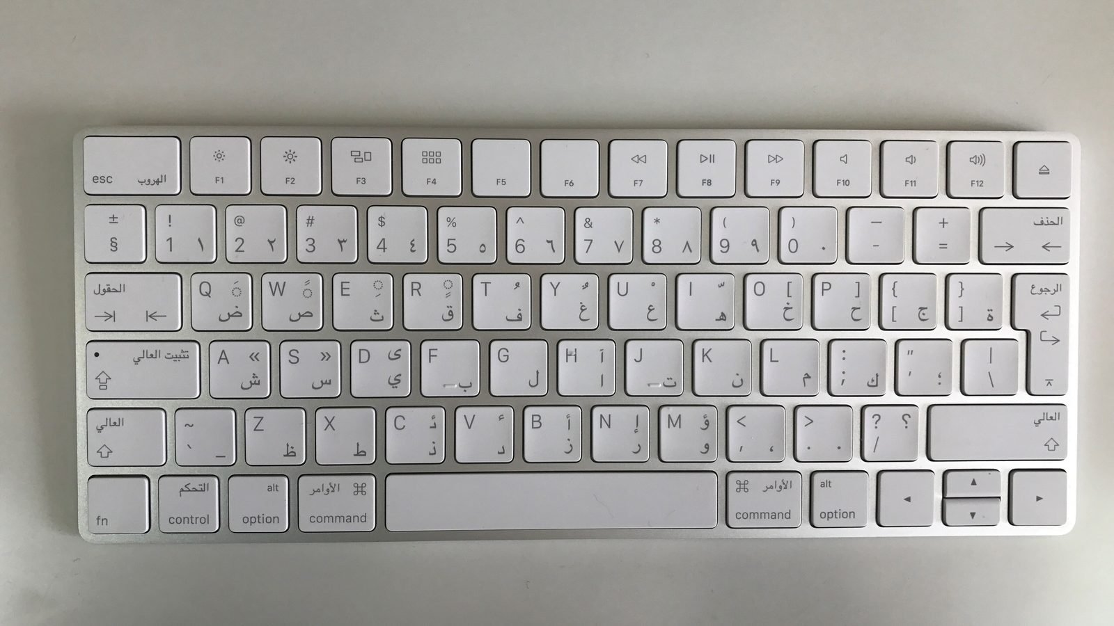 A white wireless keyboard with Arabic and English letters on each key, seen from above on a grey surface
