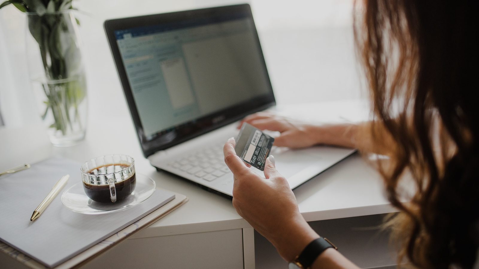 A woman holding a payment card beside her laptop while paying for an online order, face turned away