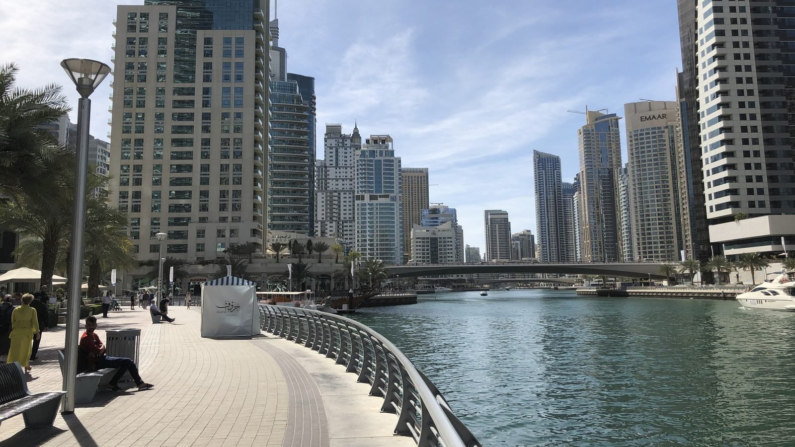 People strolling and sitting on benches along the Dubai Marina promenade, with residential towers on both sides of the water