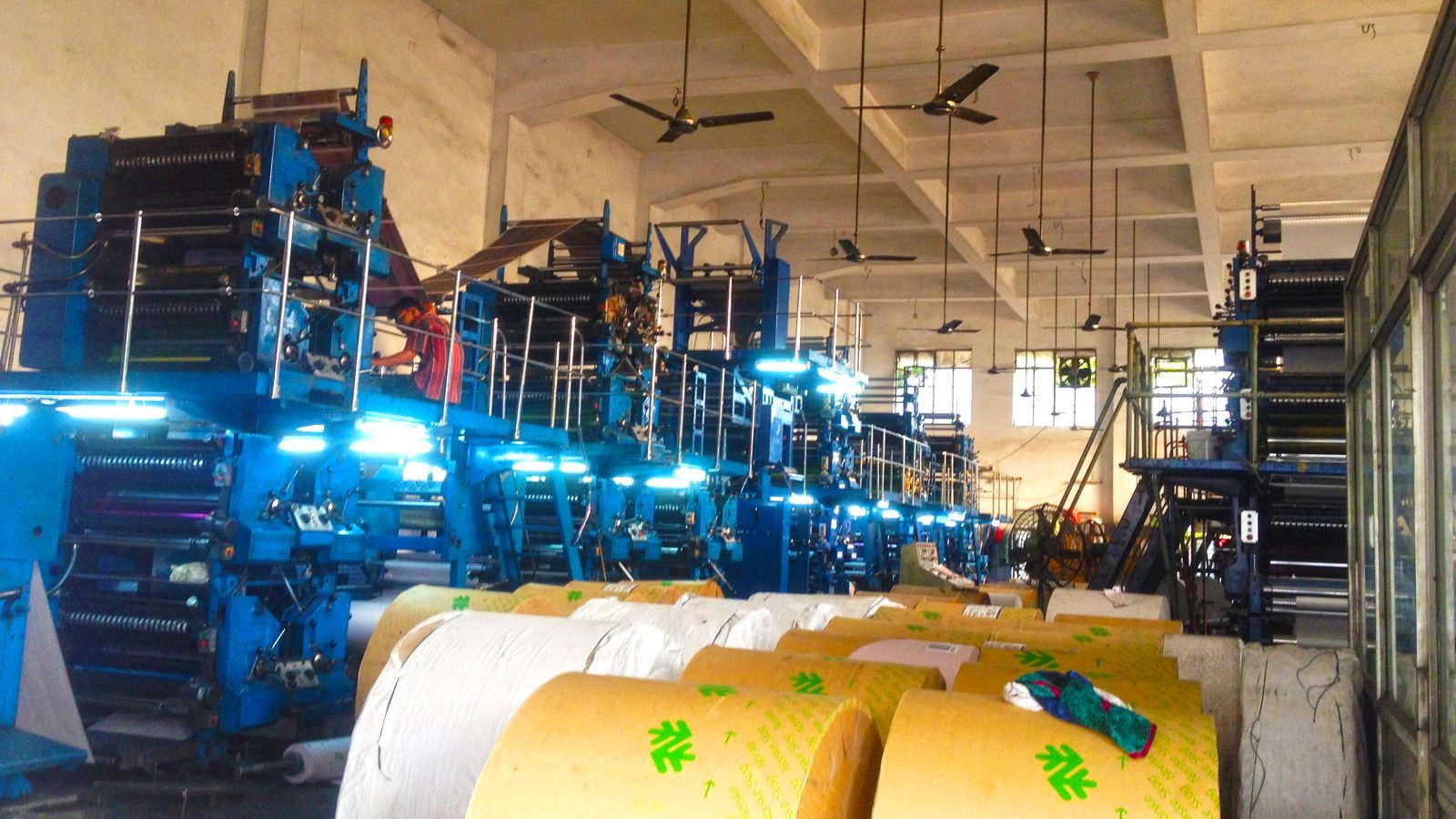 A row of blue-lit offset printing presses in a print hall, with large reels of paper stock in the foreground