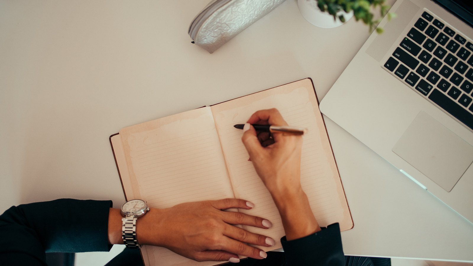 Hands writing in a notebook beside a laptop on a pale desk