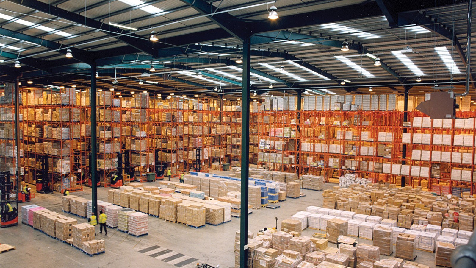 A large warehouse with tall orange pallet racking and pallets stacked on the floor